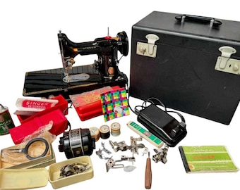 Puede incluir: Una máquina de coser Singer vintage con accesorios, que incluyen un maletín de transporte negro, un pedal y varios accesorios. La máquina es negra con detalles dorados, y hay carretes de hilo, aceite y un manual.