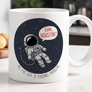 Könnte beinhalten: Ein weißer Keramikbecher mit einer kreisförmigen Grafik eines im Weltraum schwebenden Astronauten. Der Text in einer Sprechblase lautet "ERR, HOUSTON?" und der Text unter dem Astronauten lautet "ALL I'VE GOT IS FUCKING PROBLEMS" in Schwarz.