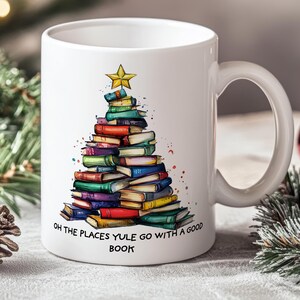 Peut inclure: Mug en céramique blanche avec un motif d'arbre de Noël fait de livres colorés, surmonté d'une étoile dorée. Le mug porte l'inscription "OH THE PLACES YULE GO WITH A GOOD BOOK".