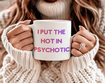 May include: White ceramic mug with the text "I PUT THE HOT IN PSYCHOTIC" in pink and purple. The mug is held by a person wearing a cream-coloured sweater.