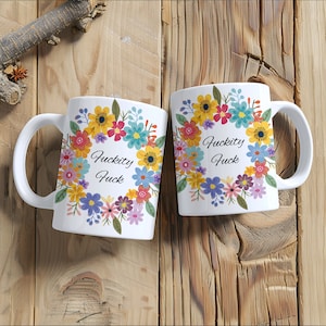 May include: Two white coffee mugs with a floral design and the text "F*ckity F*ck" on each mug. The mugs are on a wooden surface.