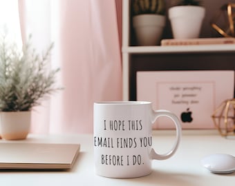 Könnte beinhalten: Weiße Keramik-Tasse mit dem Text "I HOPE THIS EMAIL FINDS YOU BEFORE I DO." Die Tasse steht auf einem weißen Schreibtisch mit einem Laptop, einer Computermaus und einem rosa Vorhang im Hintergrund. Es gibt auch Pflanzen und dekorative Gegenstände.