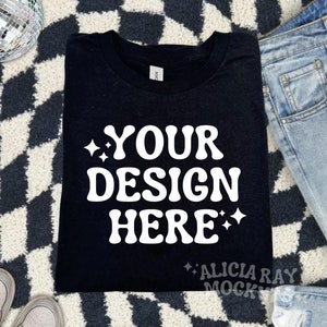 May include: A black t-shirt with the text "YOUR DESIGN HERE" in white, stylized lettering. The shirt is folded and placed on a black and white checkered rug, with a pair of light-wash jeans and sneakers visible.