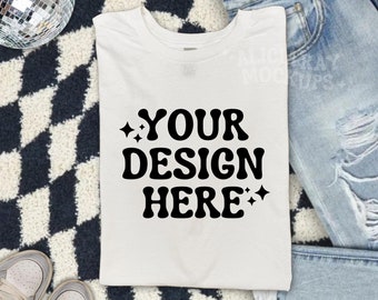 Op de afbeelding: Een wit T-shirt met de tekst "YOUR DESIGN HERE" in zwarte letters met kleine sterren aan beide kanten van de tekst. Het T-shirt ligt op een zwart-wit geblokt oppervlak met een paar witte sneakers en een blauwe jeans.