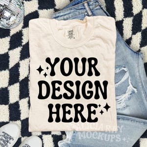 May include: Cream-colored t-shirt with the text "YOUR DESIGN HERE" in bold, black, retro-style font. The shirt is folded and placed on a black and white checkered surface, with a pair of jeans and sneakers in the background.