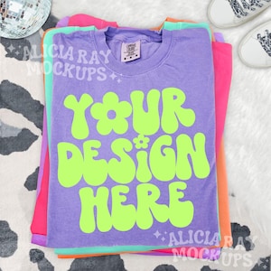 May include: A stack of colorful t-shirts in purple, pink, orange, and mint green. The top shirt is purple with the text "YOUR DESIGN HERE" in bright green. The shirts are on a white and black cow print surface.