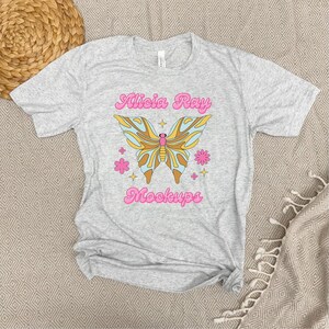 Pode incluir: Uma camiseta cinza mesclada com um desenho de borboleta e o texto "Alicia Ray Mockups". A borboleta tem asas amarelas, azuis e marrons, com flores e estrelas rosas. O texto é rosa.