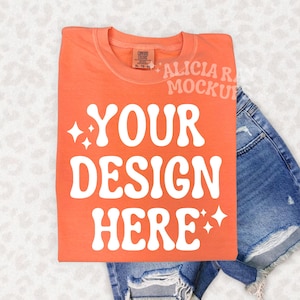 May include: An orange t-shirt with the text "YOUR DESIGN HERE" in white, with starburst accents. The shirt is folded and placed on top of a pair of denim shorts. The shirt is a Comfort Colors XL-TG-PG.