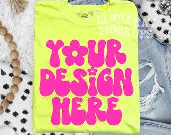 Op de afbeelding: Een neongeel t-shirt met de tekst "YOUR DESIGN HERE" in felroze, retro-stijl letters. Het shirt is opgevouwen en geplaatst op een witte, zachte ondergrond, met een spijkerbroek op de achtergrond. Decoratieve elementen zijn een discobal en houten kralen.
