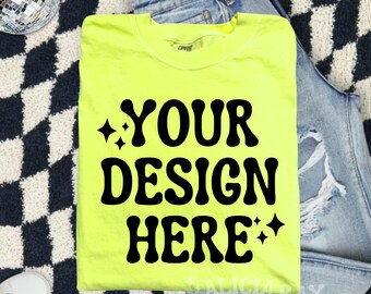 Op de afbeelding: Een felgeel T-shirt met de tekst "YOUR DESIGN HERE" in zwart. Het shirt is gevouwen en ligt op een zwart-wit geblokt oppervlak met een blauwe spijkerbroek.