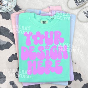 May include: A stack of Comfort Colors t-shirts in pastel colors: mint green, pink, blue, and lavender. The mint green shirt has the text "YOUR DESIGN HERE" in a retro, bubble font. A disco ball and white sneakers are in the background.