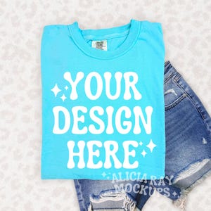 Puede incluir: Una camiseta azul claro con el texto "YOUR DESIGN HERE" en blanco. La camiseta está sobre un pantalón corto de mezclilla azul claro con detalles desgastados.