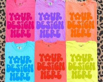 May include: Six colorful t-shirts in pink, purple, orange, blue, red, and yellow. Each shirt features the text "YOUR DESIGN HERE" in a retro, groovy font. The shirts are displayed on a leopard print background.