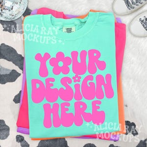 May include: A stack of folded t-shirts in shades of teal, pink, orange, and purple. The top teal shirt features the text "YOUR DESIGN HERE" in bright pink, with a flower design. The shirts are on a cow print surface.