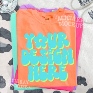 May include: An orange t-shirt with the text "YOUR DESIGN HERE" in turquoise with a yellow outline. The shirt is stacked with other t-shirts in shades of pink and green. A disco ball and white sneakers are in the background.