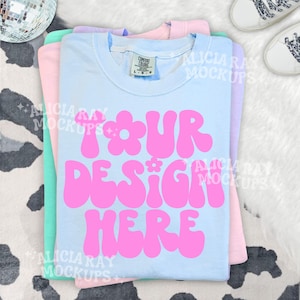 May include: A stack of pastel-colored t-shirts, including light blue, pink, and mint green. The top shirt is light blue with the text "YOUR DESIGN HERE" in pink, with a flower design. A pair of white sneakers with patterned laces are in the background.