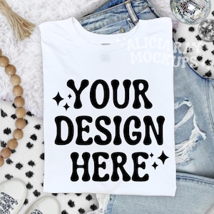 Pode incluir: Uma camiseta branca com o texto "YOUR DESIGN HERE" em negrito, preto, com uma fonte retrô. A camiseta é exibida em uma superfície macia e branca, com jeans azuis ao fundo. Inclui contas de madeira e uma bola de discoteca.