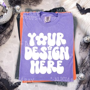 May include: A purple short-sleeved t-shirt with a white graphic print that reads "YOUR DESIGN HERE" in a bold, rounded font with flower accents. The shirt features a crew neckline and a tag indicating size M, layered over orange and dark gray shirts.