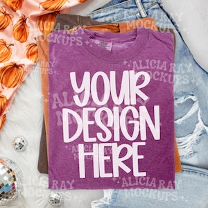 May include: A purple short-sleeved t-shirt with a crew neckline featuring the white text YOUR DESIGN HERE printed in a bold, rounded font. The shirt is layered over a brown t-shirt, a light blue denim garment, and a peach fabric with pumpkin prints.