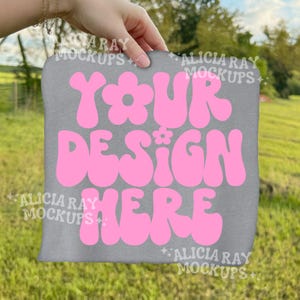 Puede incluir: Maqueta de camiseta gris con el texto "YOUR DESIGN HERE" en letras rosas estilo burbuja. La camiseta se sostiene sobre un fondo verde y herboso. Las palabras "ALICIA RAY MOCKUPS" están en marca de agua en la imagen.