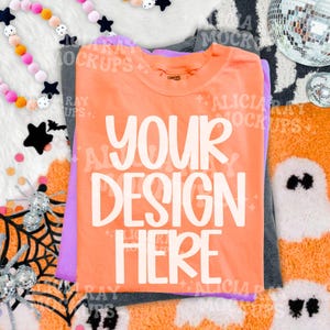 May include: A stack of three t-shirts in orange, purple, and dark gray. The top orange t-shirt features the white text "YOUR DESIGN HERE" printed in a bold, rounded font. The shirts are layered on a surface with a Halloween-themed pattern.