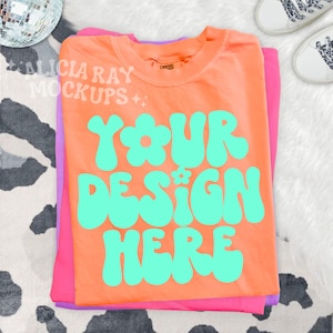 May include: An orange t-shirt with the text "YOUR DESIGN HERE" in turquoise, stacked on top of a pink and purple t-shirt. The shirts are on a white surface with a black and white cow print pattern. A disco ball and white sneakers are in the background.