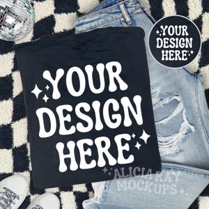 May include: Black t-shirt mockup with the text "YOUR DESIGN HERE" in white. The t-shirt is lying on a black and white chequered surface with a pair of blue jeans and a disco ball.