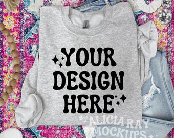 Op de afbeelding: Een grijs sweatshirt met de tekst "YOUR DESIGN HERE" in zwarte letters. Het sweatshirt ligt op een kleurrijk tapijt met een spijkerbroek en andere accessoires.