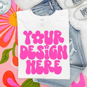 May include: White t-shirt with the text "YOUR DESIGN HERE" in bright pink, set against a backdrop of blue jeans, a colorful floral pattern, and white sneakers. The design is in a retro, groovy font.