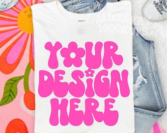 Op de afbeelding: Wit t-shirt met de tekst "YOUR DESIGN HERE" in felroze. Het shirt ligt op een blauwe spijkerbroek, met een kleurrijk bloemenpatroon en witte sneakers. De tekst is in een retro, groovy lettertype.