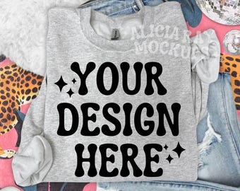 Op de afbeelding: Een lichtgrijs sweatshirt met de tekst "YOUR DESIGN HERE" in grote, zwarte letters in retrostijl. De afbeelding bevat een spijkerbroek, luipaardprint en een discobal, wat wijst op een casual, trendy thema.