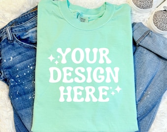 May include: A light green t-shirt with the text "YOUR DESIGN HERE" in white. The shirt is folded and laid on a white fuzzy surface with blue jeans.