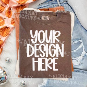 May include: A brown short-sleeve t-shirt with a crew neckline and the white text YOUR DESIGN HERE printed on the front. The shirt features a Comfort Colors brand label at the inner neck and is layered with other fabric items in a flat lay arrangement.