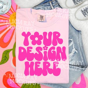 May include: A light pink t-shirt with the words "YOUR DESIGN HERE" in bright pink, stylized font. The shirt is laid on top of a pair of blue jeans. White sneakers are visible in the background.