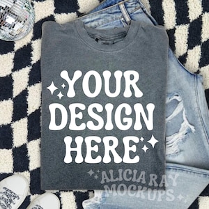 May include: A gray t-shirt with the text "YOUR DESIGN HERE" in white. The shirt is laying on a black and white checkered surface with a pair of blue jeans and a disco ball.