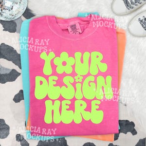 May include: A stack of three t-shirts in bright colors: pink, orange, and turquoise. The pink shirt features the text "YOUR DESIGN HERE" in a retro, lime green font. The shirts are neatly folded, showcasing a mock-up design.