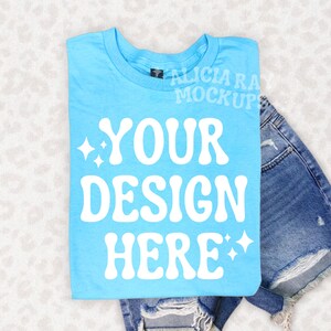 Peut inclure: Un t-shirt bleu clair plié avec le texte blanc "YOUR DESIGN HERE". Le t-shirt est présenté avec un short en jean. L'image est une maquette, parfaite pour présenter des créations personnalisées sur des vêtements.