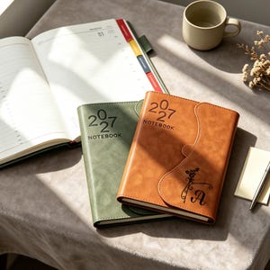 May include: Two notebooks with leather-effect covers, one olive green and one tan, both embossed with 2027 NOTEBOOK. The tan notebook features a curved flap closure, a rose illustration, and the letter A. An open planner with coloured tabs is visible.