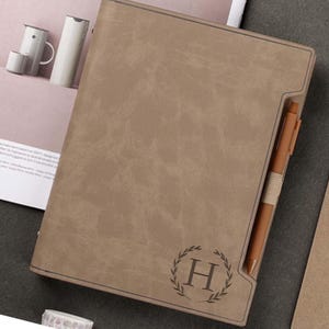 May include: A brown leather-bound journal with a pen holder and a personalised monogram of the letter "H" surrounded by a laurel wreath.
