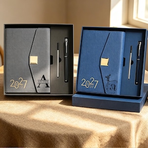 Könnte beinhalten: Zwei Geschenksets mit einem Notizbuch mit goldenem quadratischem Verschluss, einem Stift und einer Mine. Das graue Notizbuch zeigt den Text 2027 und Archer mit einem A. Das blaue Notizbuch zeigt 2027, eine Rosenillustration und den Buchstaben A.