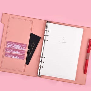 May include: A pink leather bound notebook with a silver ring binder. The notebook has a pocket on the front cover and a card holder on the inside. The notebook is open to a blank page with the word "Remember" written in small font at the top of the page.