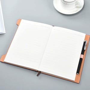 May include: A brown leather-bound journal with lined pages and a pen holder. The journal is open to a blank page.