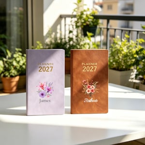 May include: Two rectangular planners for the year 2027. The left planner is light purple with a floral graphic and the name James. The right planner is brown with a floral graphic and the name Arthur. Both feature gold text reading PLANNER 2027.