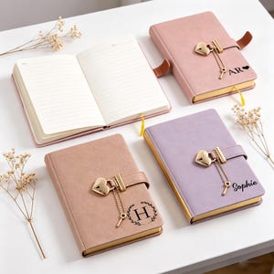 Könnte beinhalten: Vier ledergebundene Notizbücher in Rosa, Mauve und Lavendel, mit goldfarbenen Seiten. Jedes Notizbuch hat einen herzförmigen Schlossverschluss mit einem Schlüsselanhänger. Ein Notizbuch ist geöffnet und zeigt linierte Seiten. Die Notizbücher sind mit Initialen und Namen personalisiert.