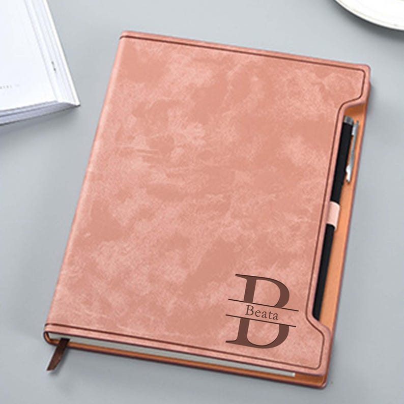 May include: A blush pink leather-bound journal with a brown leather pen holder and a personalized inscription that reads "B  Beata".