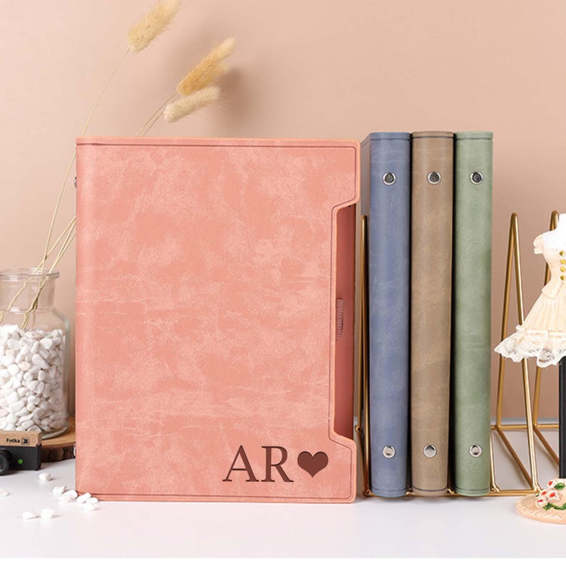 May include: Four binders with different coloured covers. The binder on the left is pink with the initials "AR" and a heart. The other binders are blue, brown, and green.