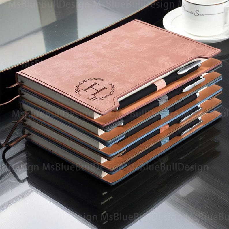 May include: A stack of four brown leather journals with a personalized monogram of the letter "H" in a wreath design. The journals have blue and brown accents and are stacked on a black surface.
