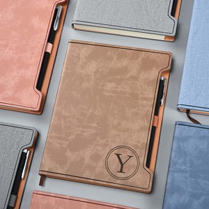 May include: A set of five notebooks with different colored covers. The notebooks have a textured, leather-like finish. The notebook in the center has a brown cover with a circle containing the letter "Y" in the center. Each notebook has a pen holder on the side.