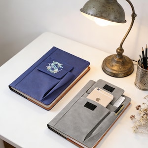 May include: Two notebooks with pockets. One is navy blue with a floral design on the pocket, the other is gray. The gray notebook has a smartphone and pen inside the pocket. Both notebooks have a brown border.