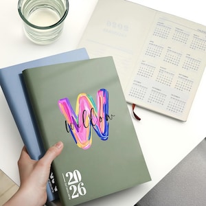 May include: A green planner with the word "willow" in colourful brushstrokes and the numbers "20 26" printed on the cover. A blue planner and an open planner with a calendar are also visible.